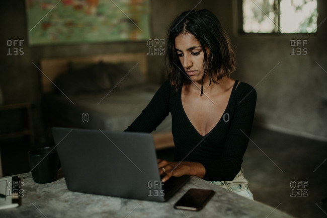 Serious focused youthful ethnic female typing on laptop while sitting at table on table kitchen modern marble countertop