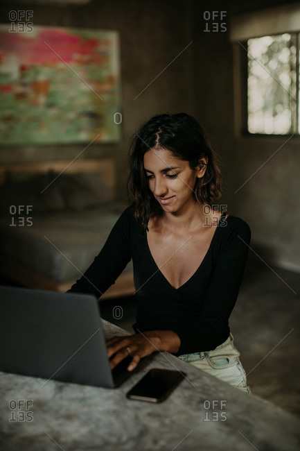 Serious focused youthful ethnic female typing on laptop while sitting at table on table kitchen modern marble countertop