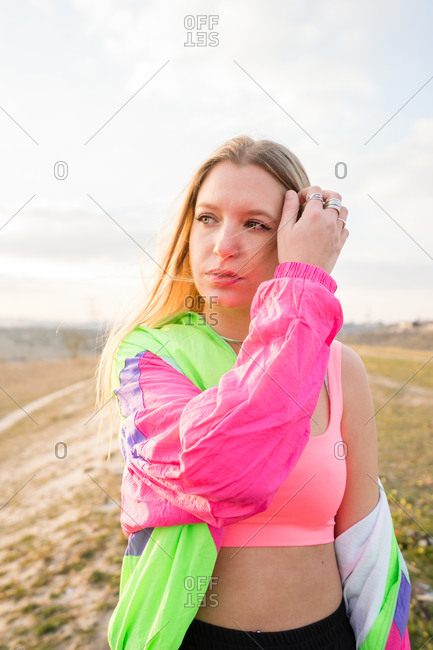 Contemporary blond haired young woman in pink bra with colorful jacket looking away with remote land on background