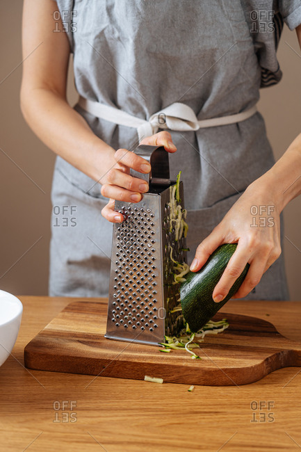 Cropped female in grey apron grating fresh zucchini while standing at wooden table in kitchen and preparing healthy dinner