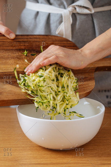 Cropped housewife putting grating zucchini from wooden cutting board into large white bowl while preparing food an wooden table