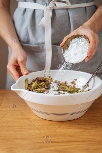 Cropped female adding salt into white bowl with mixed ingredients while preparing dinner at wooden table in kitchen
