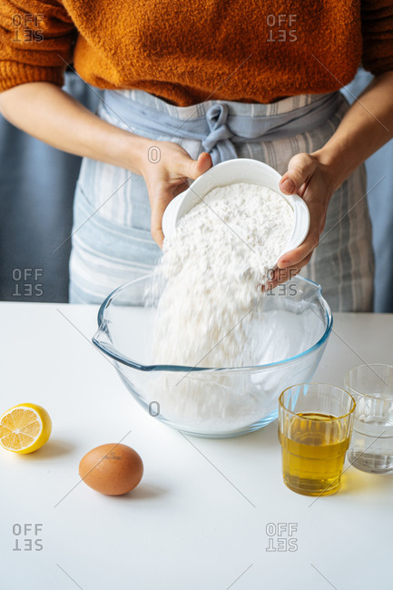 Cropped housewife putting white flour into large glass bowl while preparing dough at white table with prepared ingredients