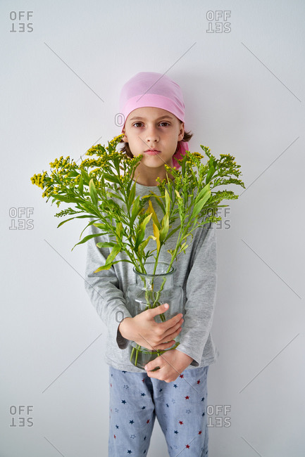 Focused little boy with cancer diagnosis wearing pink bandana and looking at camera while holding vase with flowers and standing at wall