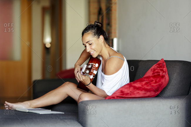 Middle aged woman is composing music with her guitar at home on the sofa.