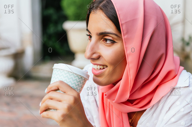 Portrait of a beautiful young Muslim woman with pink hijab smiling and drinking take-away coffee in a garden