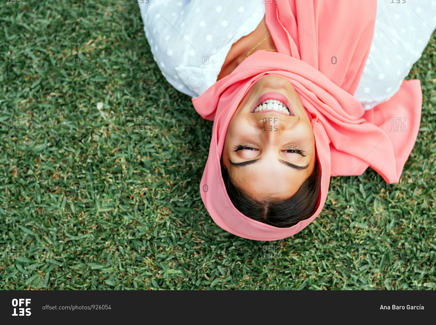 Portrait of a beautiful young Muslim woman with pink hijab lying on her back and smiling on the green grass