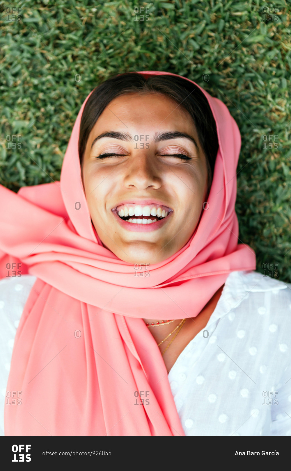 Portrait of a beautiful young Muslim woman with pink hijab lying on her back and laughing with eyes closed on the green grass