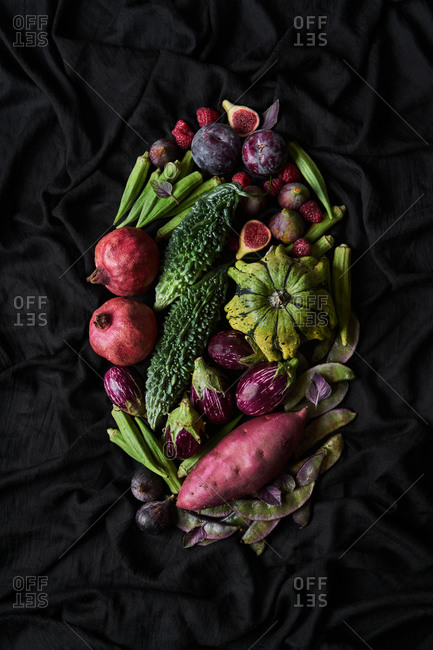 Top view of basket with assorted colorful fresh fruits and vegetables including pomegranate and plums and figs with mini eggplants and sweet potato on black background