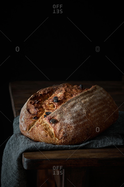 Crispy freshly baked sourdough bread loaf with raisin placed on gray cloth on wooden table with black background