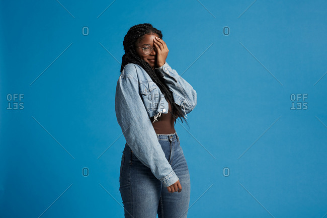 Plus size black female with braids in casual clothes smiling looking at camera covering face with hand against blue background