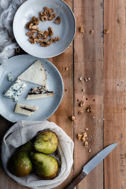 From above cotton sack with fresh pears and plate with walnuts placed near cheese and knife on timber table