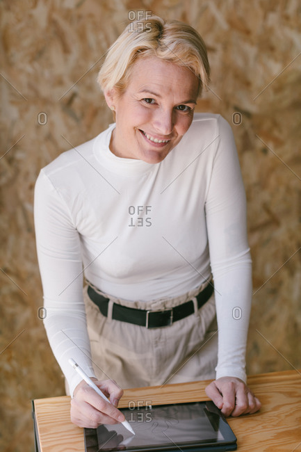 Stylish blond businesswoman smiling looking at camera while bending over table and working on tablet with stylus in light wooden office