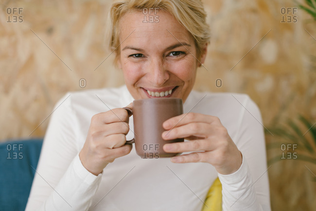 Calm adult businesswoman with short blonde hair sitting on cozy sofa in office having mug of coffee and smiling calmly at camera
