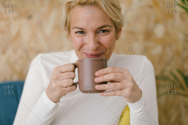 Calm adult businesswoman with short blonde hair sitting on cozy sofa in office having mug of coffee and smiling calmly at camera