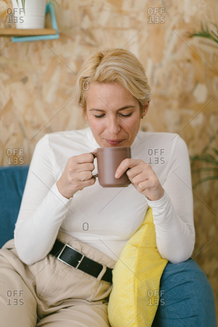 Calm adult businesswoman with short blonde hair sitting on cozy sofa in office having mug of coffee and smiling calmly looking away