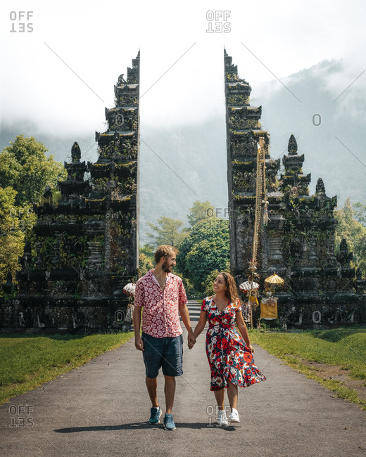 Happy young couple on vacation in light casual wear holding hands smiling and looking at each other while walking on empty path among exotic ancient constructions in Indonesia