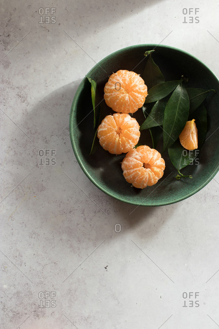 Top view of green ceramic bowl with fresh peeled tangerines placed on white table near unpeeled fruits with green leaves