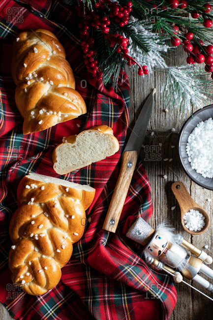 Top view of wooden table with cut traditional braided bread and knife placed on checkered Christmas tablecloth with decorative objects