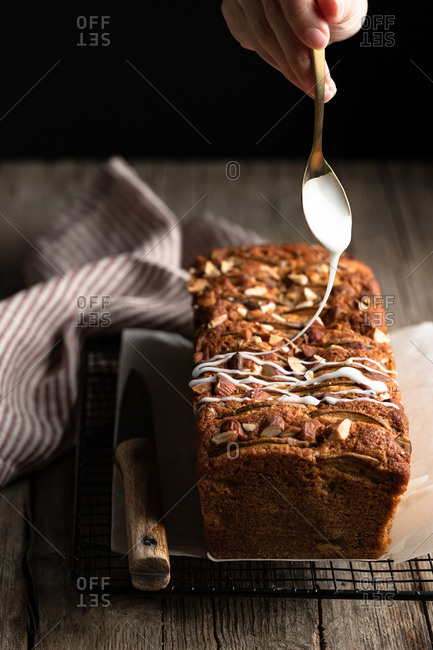 Crop housewife with spoon in hand pouring white sugar icing over homemade banana bread with nuts placed on metal grid on wooden table