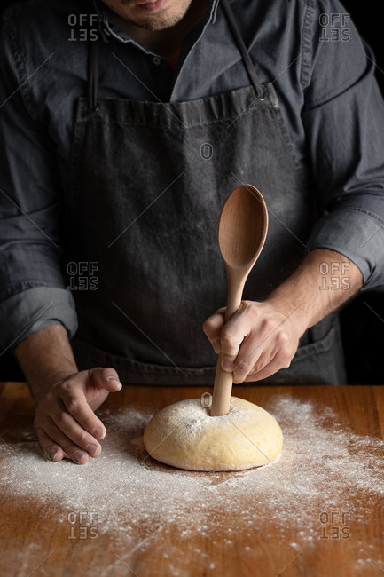 Crop male baker in black apron using big wooden spoon for making hole in dough while forming artisan round bread loaf at wooden table