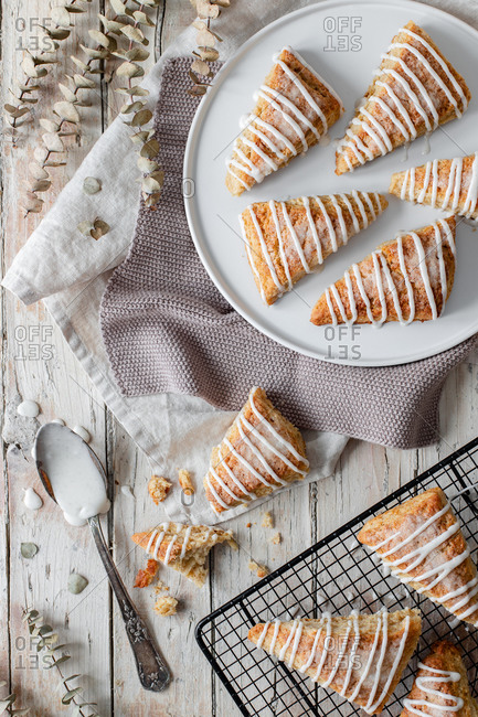 Top view of tasty sweet homemade triangle scones decorated with sugar icing placed on white plate and on metal grid on wooden table with decorative tree cones