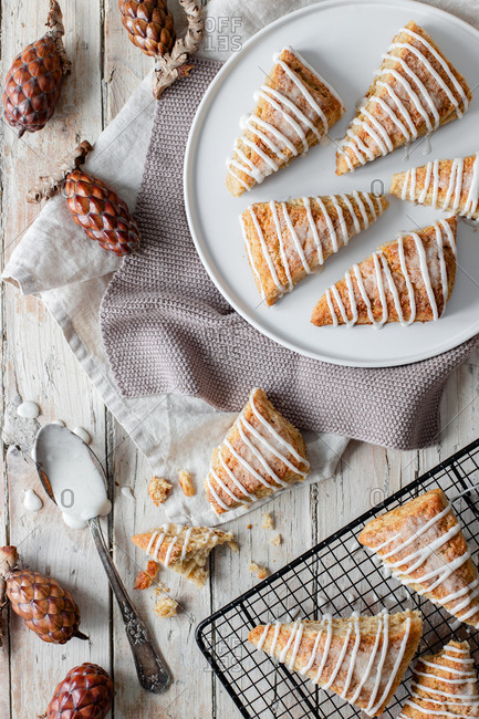 Top view of tasty sweet homemade triangle scones decorated with sugar icing placed on white plate and on metal grid on wooden table with decorative tree cones