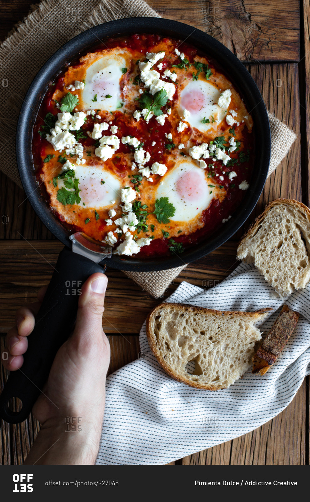 Top view of crop person putting on wooden table frying pan with delicious shakshuka made with fried eggs with vegetables and feta cheese
