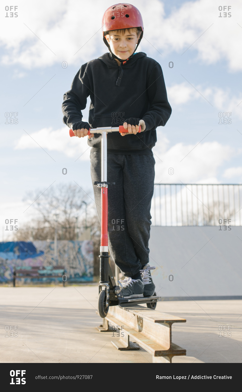 Active teen boy in protective helmet with kick scooter standing on ramp in skate park while preparing for performing trick in sunny spring day