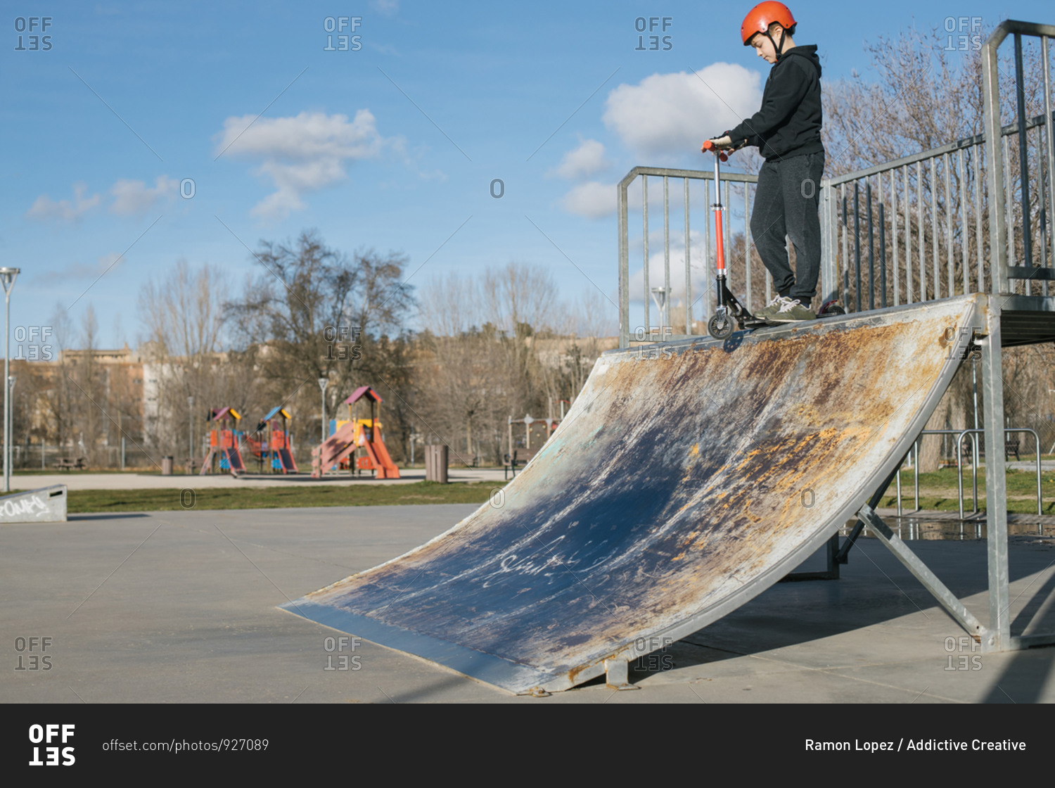 From below of active teen boy in protective helmet with kick scooter standing on ramp in skate park while preparing for performing trick in sunny spring day