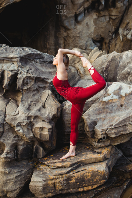 Side view of young woman in red sportswear doing yoga on lord of the dance pose standing on one leg while outstretching different leg on rough rock among mountains