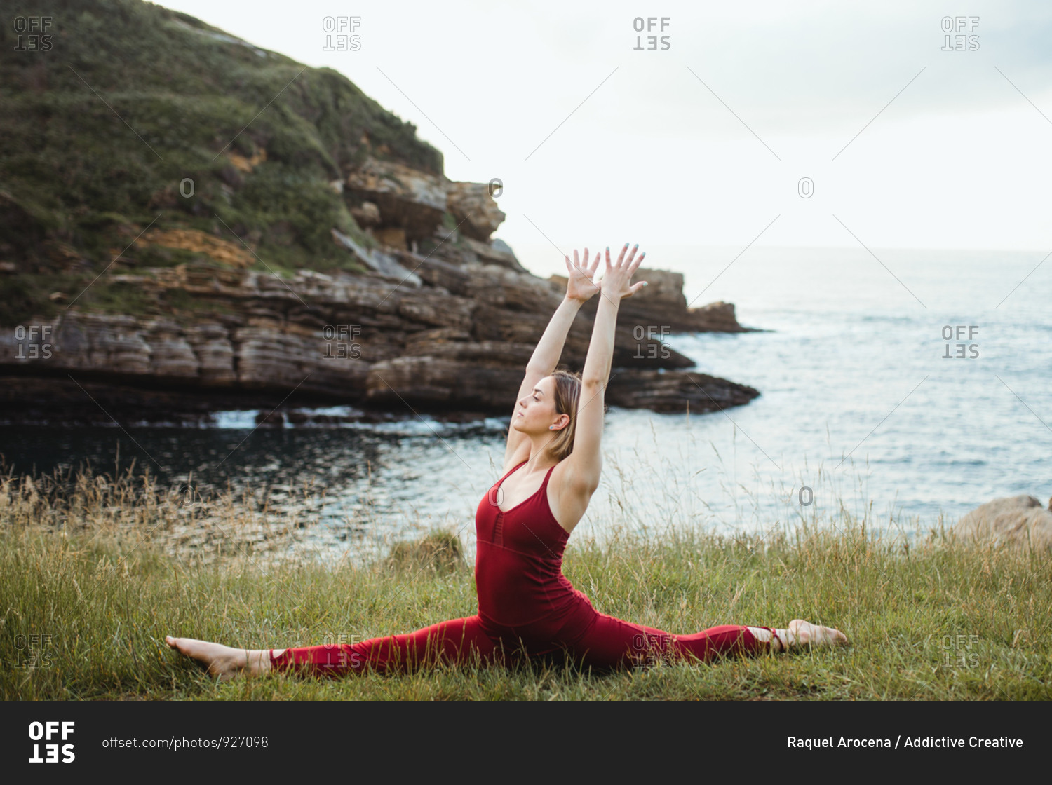 Young flexible woman in red sportswear sitting in difficult yoga monkey pose on empty green hill
