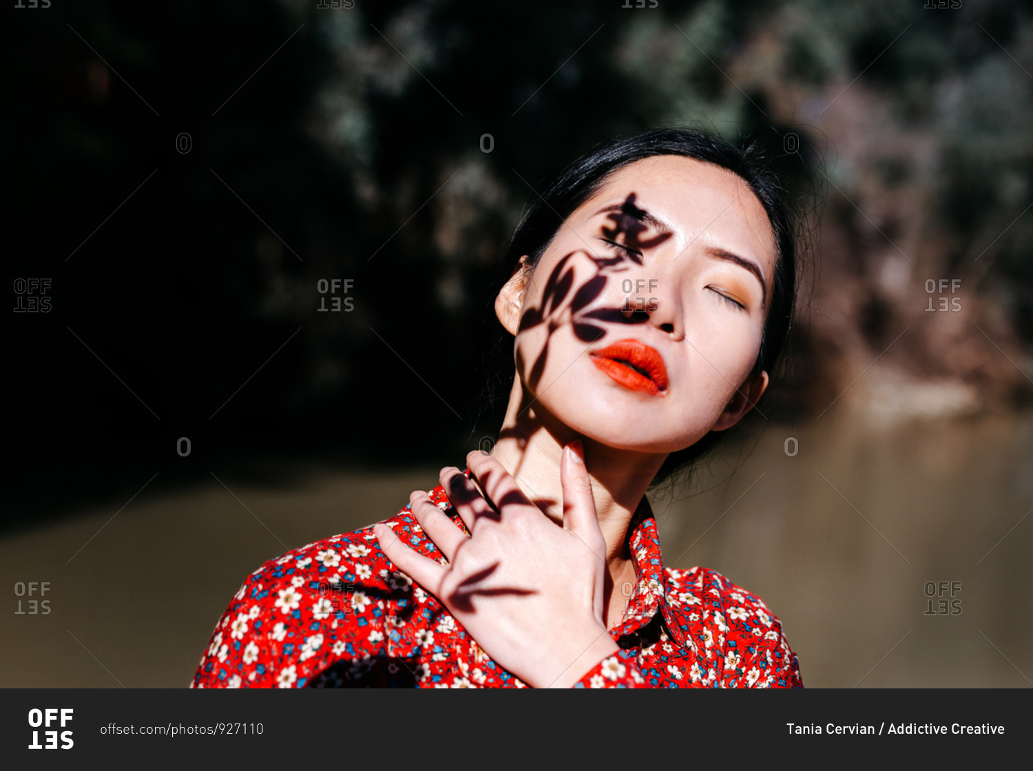 Beautiful Asian lady with closed eyes and shadow of plant twig on face standing against lake in countryside
