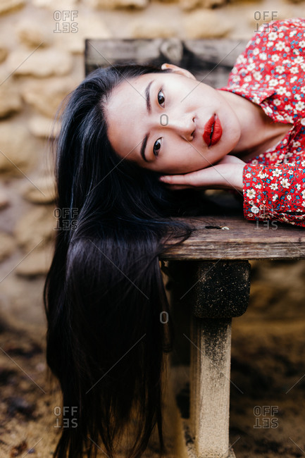 Pretty ethnic lady in trendy floral clothes lying on old lumber bench and looking at camera against stone wall