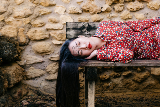 Pretty ethnic lady in trendy floral clothes lying on old lumber bench with closed eyes against stone wall