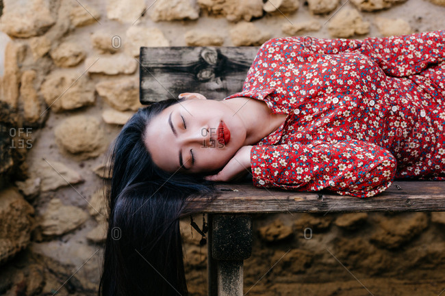 Pretty ethnic lady in trendy floral clothes lying on old lumber bench with closed eyes against stone wall