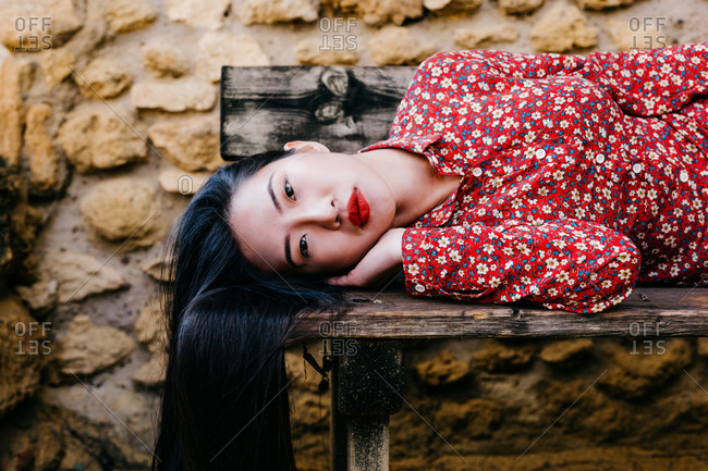 Pretty ethnic lady in trendy floral clothes lying on old lumber bench and looking at camera against stone wall