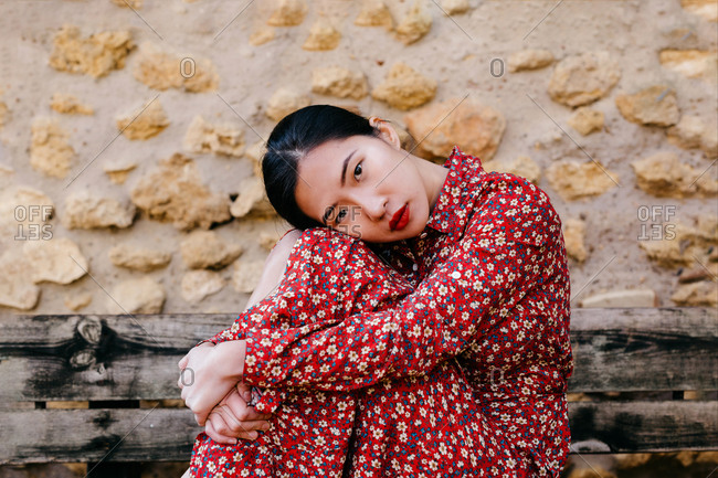 Side view of attractive Asian lady in floral dress embracing knees and looking at camera while sitting on shabby wooden bench against stone wall