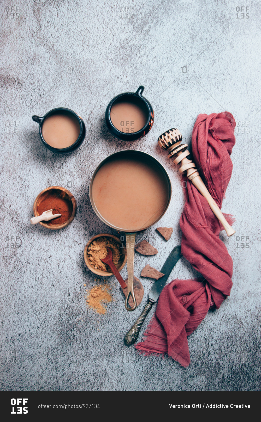 Top view of traditional Mexican spicy hot chocolate with cinnamon and red pepper powders served in pot and cups placed with chunks of chocolate and wooden whisk with red cloth on gray background