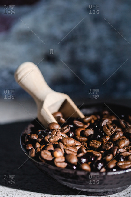 From above of black bowl with aromatic fresh roasted coffee beans and wooden serving scoop placed on table with blurred background