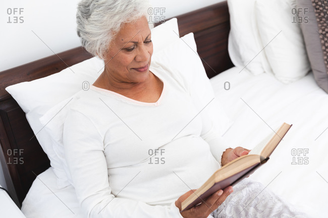 Close up of an attractive retired senior African American woman at home, sitting up in bed reading a book and smiling, self isolating during coronavirus covid19 pandemic