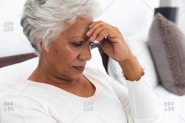 Close up of a senior retired African American woman at home sitting up in bed in her bedroom with a headache, touching her head and looking down, self isolating during coronavirus covid19 pandemic