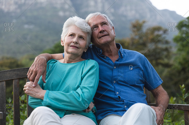 Happy retired senior Caucasian couple at home in the garden outside their house, sitting on a bench, relaxing and embracing, both looking away and smiling, at home together isolating during coronavirus covid19 pandemic