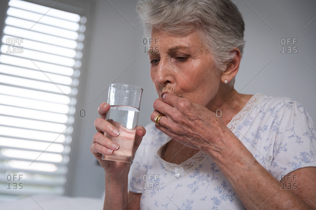 Close up of a retired senior Caucasian woman at home sitting up in bed in her bedroom holding a glass of water and taking medication, self isolating during coronavirus covid19 pandemic
