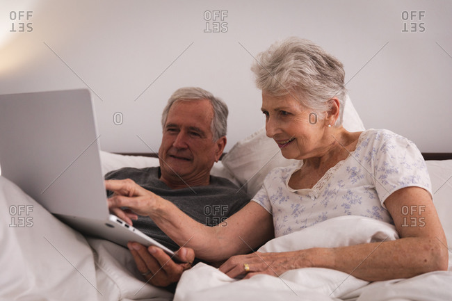 Close up of a happy retired senior Caucasian couple at home sitting up in bed in their bedroom, talking and using a laptop computer together and smiling, couple isolating during coronavirus covid19 pandemic
