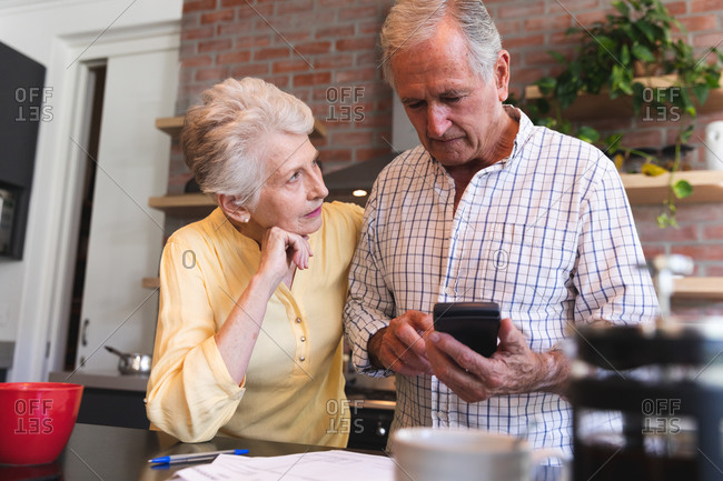 A retired senior Caucasian couple standing at a table in their dining room using a calculator and discussing their finances, with a pot of coffee and cup on the table in the foreground, at home together isolating during coronavirus covid19 pandemic