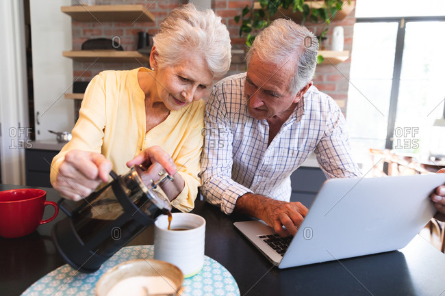 A retired senior Caucasian couple at home standing at a table in their kitchen, talking and smiling, using a laptop computer together and smiling, the woman pouring coffee from a pot, couple isolating during coronavirus covid19 pandemic