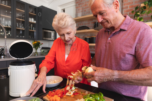 Happy retired senior Caucasian couple at home, preparing food and smiling in their kitchen, the man cutting vegetables, the woman watching and talking to him, at home together isolating during coronavirus covid19 pandemic