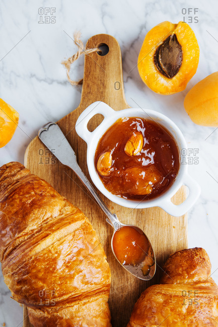 Top view of appetizing fresh croissant served with pot of homemade apricot jam on wooden cutting board placed near fresh fruits on marble background