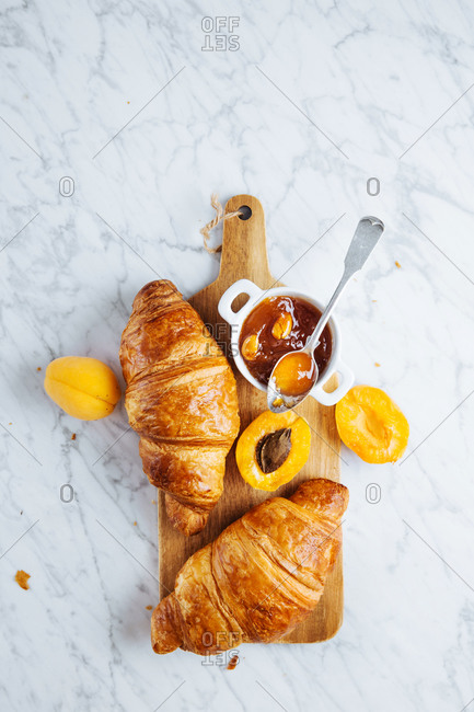 Top view of appetizing fresh croissant served with pot of homemade apricot jam on wooden cutting board placed near fresh fruits on marble background